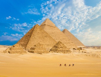 all egyptian pyramids from distance with row of camels walking in foreground in giza, cairo, egypt.  wide telephoto shot plenty of copy space.