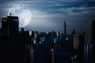 silhouettes of skyscrapers different construction in the dark town with background of a large moon and clouds at nighttime. dark tone and high contrast style. the moon were not furnished by nasa.