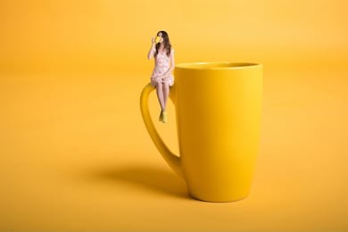 creative surrealism design with miniature people. girl with a cup. the girl in a pink dress sits on the mug. yellow cup of tea or coffee. young woman drinking tea. mug on yellow background