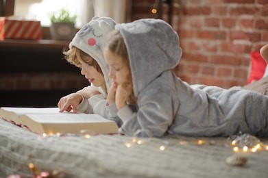 christmas holidays. two children, brother and sister lie on big bed and read christmas tales. children dressed in fluffy pajamas, like little cute animals. before them the huge book. magic christmas.