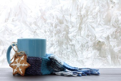 blue mug with a warm scarf and homemade cakes on the background of frozen window / drinks for the winter season