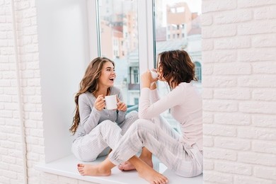 two pretty girls in pajamas sitting on window in light room. they drinking tee and smiling to city outside