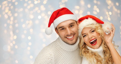 people, christmas, holidays and new year concept - happy family couple in sweaters and santa hats over holidays lights background