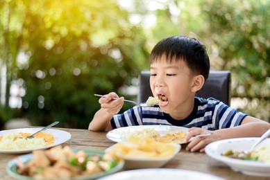 selective focus at young thai boy eating with food on the wooden table with his family in the garden. and out focus tree background.