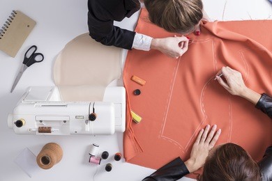 top view of two girls making a pattern on a red piece of tissue. sewing machine is standing on the table beside them.