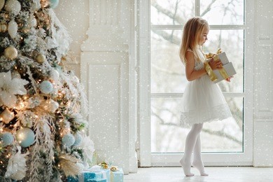 beautiful little girl standing on tiptoes at the large window and holding a gift, in the bright new year's interior