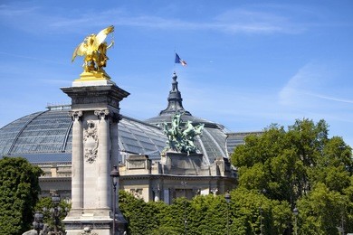 view of grand palais and golden statue at pont alexandre iii bridge. french flag waves on top of the building. trees and blue sky are in the background.