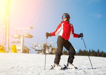 woman wearing helmet, red jacket and ski goggles standing with skis on mountain top at a winter resort in sunny day with ski lifts and blue sky in background. bukovel, ukraine