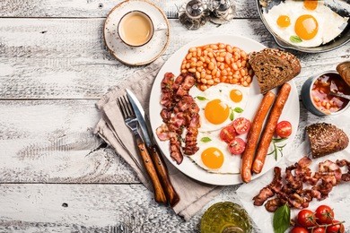 traditional english breakfast - sunny-side-up fried eggs, sausages, beans and bacon on white plate with coffee, top view