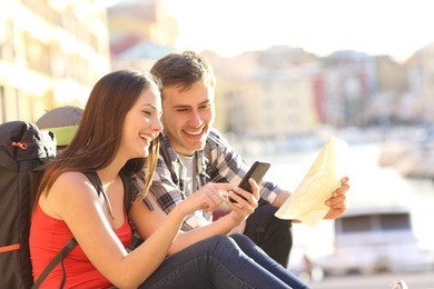two tourists searching location on a smart phone sitting on the floor of a travel destination in the coast with a warm light in the background
