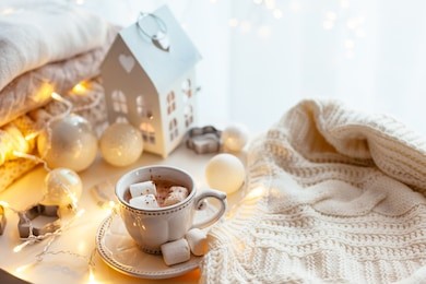 white christmas decor: warm sweater, cup of hot cocoa with marshmallow and led string lights. winter mood, holiday decoration.
