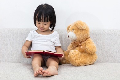 asian chinese little girl reading book with teddy bear on the sofa in the living room.