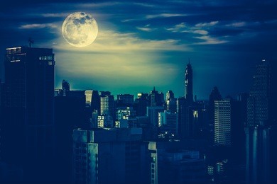 silhouettes of skyscrapers different construction in the dark town with background of a large moon and clouds at nighttime. cross process and dark tone. the moon were not furnished by nasa.