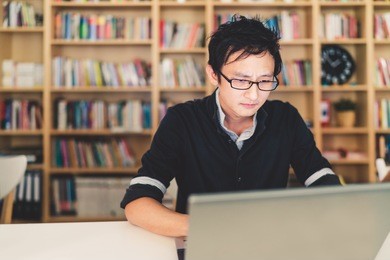 young pensive asian man working on laptop at home office or library with serious face, bookshelf with clock blur background with copy space, business or technology concept