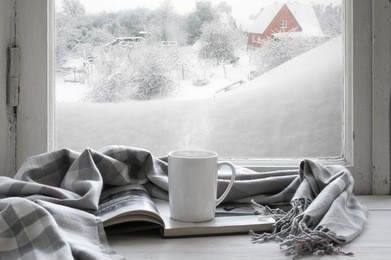 cozy winter still life: mug of hot coffee and opened book with warm plaid on vintage windowsill of cottage against snow landscape with snowdrift from outside.
