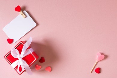 valentine day composition: red gift box with bow, credit / visiting card template with clamp, candy and small hearts on light pink background. top view.