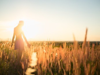 trendy girl in stylish summer dress with beautiful hat walking in the field with flowers in sunlight