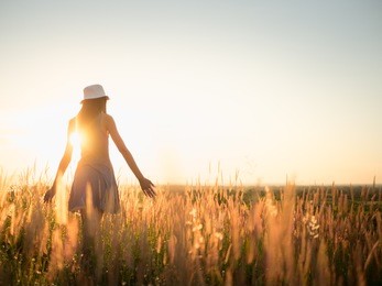 trendy girl in stylish summer dress with beautiful hat walking in the field with flowers in sunlight