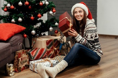 beautiful happy emotional woman in red santa hat and reindeer sweater smiling after receiving christmas presents near decorated christmas tree, greeting card concept 