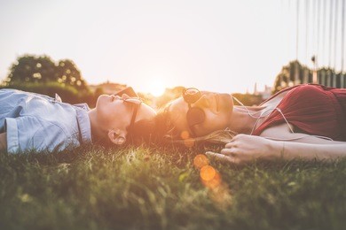 summer sunny evening. backlight. two young woman in sunglasses lying on grass heads together. one of the girls listening to music on headphones. blurred background