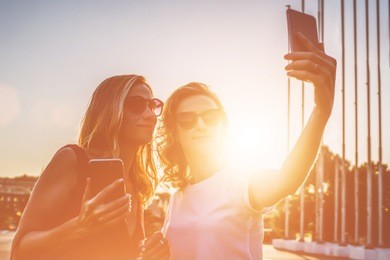 summer sunny day, backlight, two smiling young women in sunglasses with blond hair standing outside and take pictures on your smartphone. girls using a digital gadgets. film effect.instagram filter