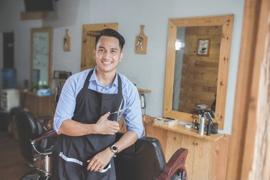 happy young business owner looking at camera while leaning on chair at his barbershop