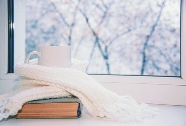 mug of hot tea and warm woolen knitting on windowsill against snow landscape from outside.
