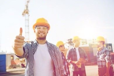 business, building, teamwork, gesture and people concept - group of smiling builders in hardhats showing thumbs up outdoors