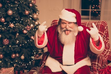 it's party! cool santa claus showing tongue out and gesturing with fingers rock-and-roll near christmas tree at home