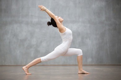 side view portrait of beautiful young woman wearing white tank top working out against grey wall, doing yoga or pilates exercise. standing in warrior one pose, virabhadrasana. full length