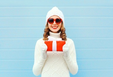 christmas happy smiling young woman with gift box wearing a knitted hat sweater sunglasses over blue background