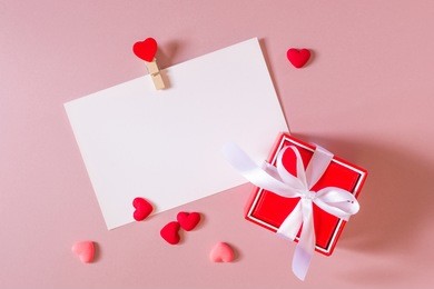 valentine day composition: red gift box with bow, stationery / photo / postcard template with clamp and small hearts on light pink background. top view.