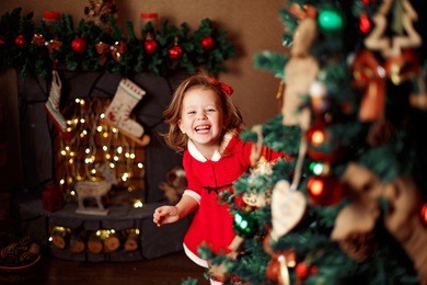smiling little girl peeping from behind christmas tree in living room. horizontal indoors shot
