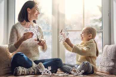 merry christmas and happy holidays! happy loving family sitting by the window and making paper snowflakes for decoration windows. mother and child creating decorations.