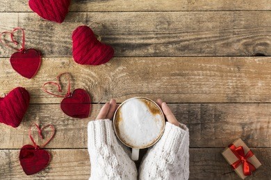 woman in white knitted sweater is holding a cup of hot cappuccino with decorative hearts around for the valentine's day