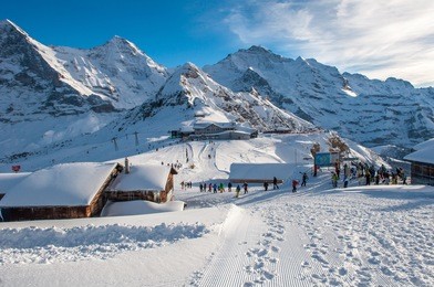 lots of people with snowboards and skis on the mountain top getting ready for snow fun. sun shining/sunlight on the snow covered wooden cottages, in engelberg, mount titlis, switzerland.  