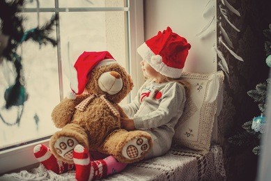 little girl in a santa claus hat with teddy bear sitting on the window. the child looks out the window and waiting for christmas, santa claus