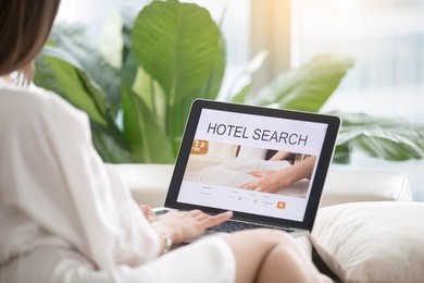 young woman wearing a dressing gown sitting comfortably with laptop searching hotel accommodations online, trying to save on hotel stays, booking a room, checking out a selection of resorts