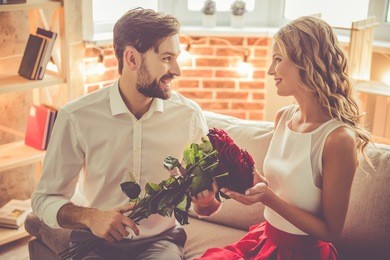 handsome elegant guy is giving his beautiful girlfriend roses and smiling while they having a romantic date at home