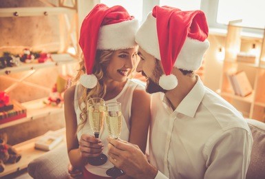 beautiful young couple in santa hats is holding glasses of champagne and smiling while celebrating new year at home