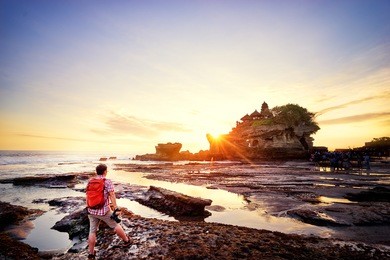 travel and photography. young man with camera taking picture of beautiful balinese landscape. ancient hinduism temple tanah lot on the rock against sunset sky. bali island, indonesia.