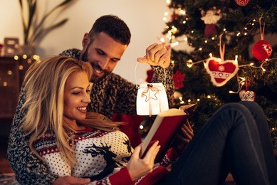 couple enjoying a book together while lying next to christmas tree.