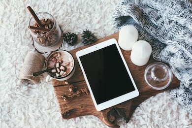 winter homely scene, scandinavian style. warm knit sweater, tablet pc with blank screen, candles, cup of sweet cocoa with marshmallows and other decor on wooden tray in bed. lazy cold weekend.