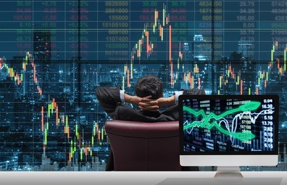 back side of sitting businessman who is looking at stock market exchange graph over the cityscape with computer show trading graph background on the table, business technology and trading concept