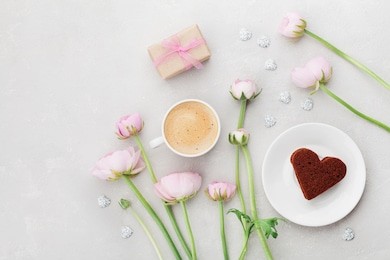 breakfast for valentines day with cup of coffee, gift, flowers and cake in shape of heart on gray table from above in flat lay style.
