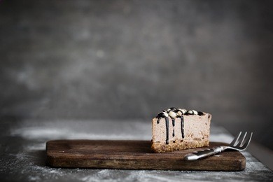 cut of raw chocolate mousse cake with cashew, hazelnuts and dark chocolate glaze topping on a wooden and grey background. vegan sugar gluten free dessert. dark food photography. copy space, horizontal