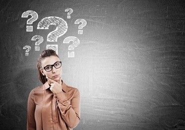 thinking woman in glasses with her fingers on the chin is standing near a blackboard with question marks. mock up
