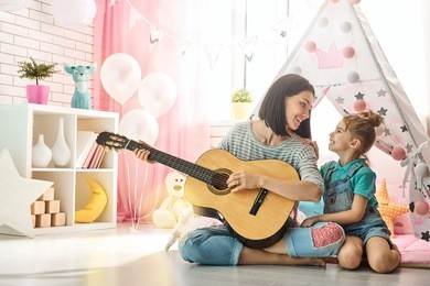 happy loving family. pretty young mother and daughter playing guitar together. adult woman playing guitar for child girl indoors. funny mom and lovely child having fun in children room.