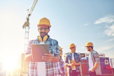 business, building, industry, technology and people concept - smiling builder in hardhat with tablet pc computer over group of builders at construction site