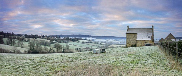 a panorama view of a cold and frosty sunrise over an isolated farm house and a distant view of the cotswold village of painswick, gloucestershire, england, uk